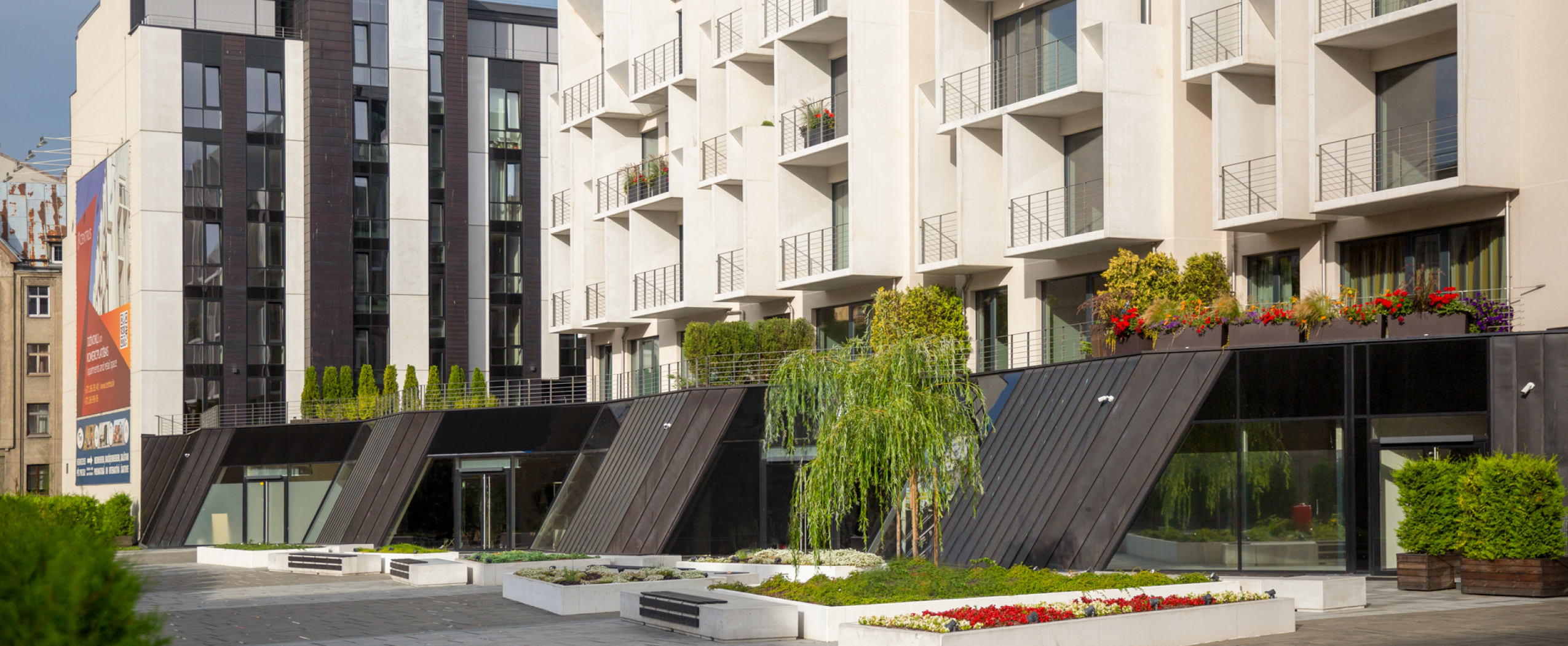 CENTRUS kvartāla promenāde ar kokiem, apstādījumiem un fonā redzamām pirmās kārtas ēkām Rīgas centrā.